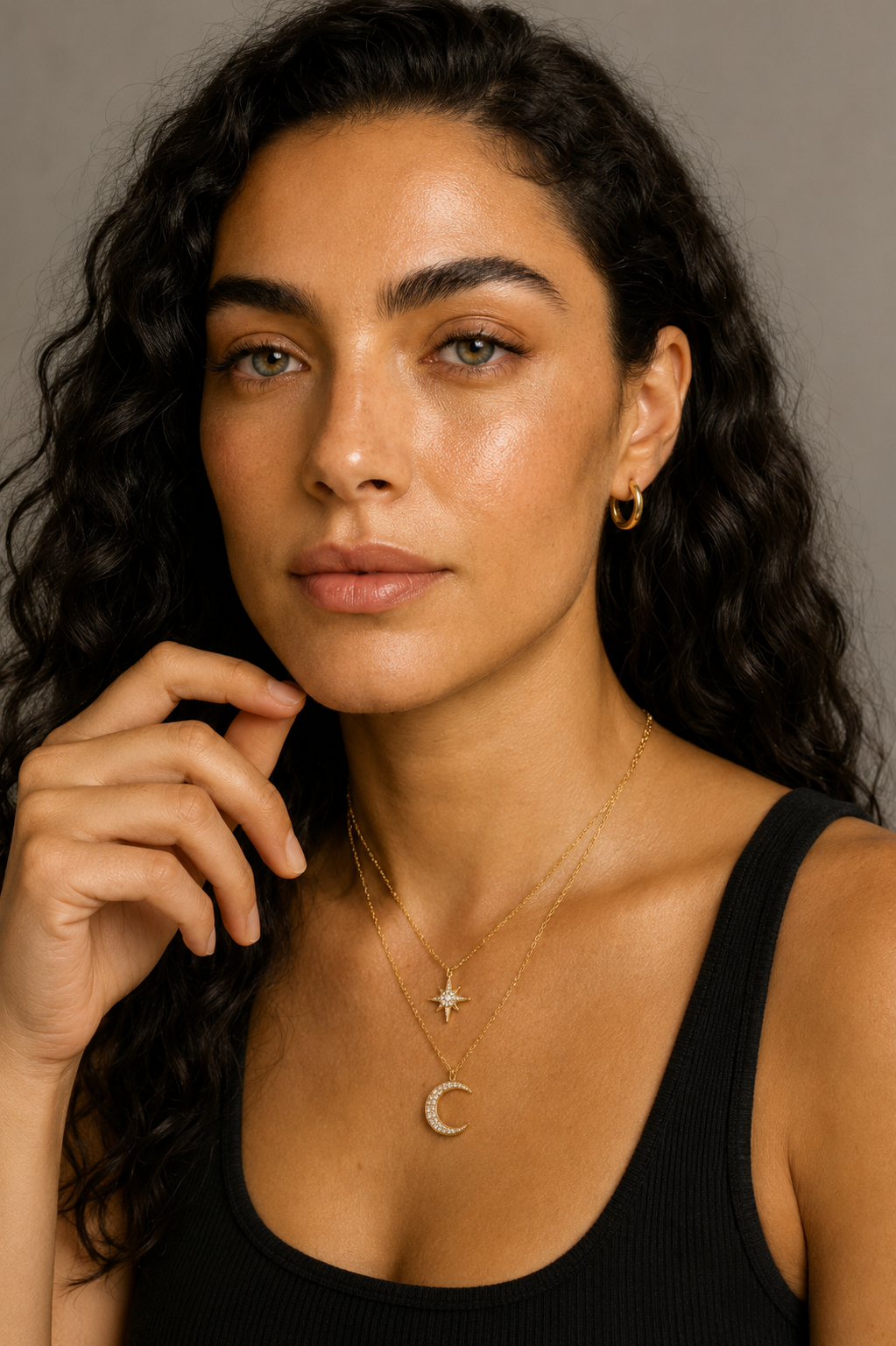 Woman wearing layered gold necklaces featuring a star pendant and crescent moon pendant with black sleeveless top against a neutral background.