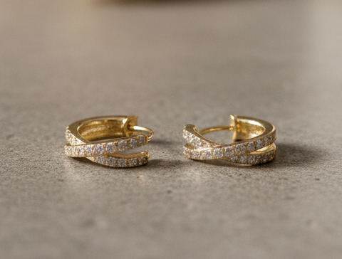 Gold crisscross huggie earrings with crystal detailing from Dione Canada displayed on a textured surface