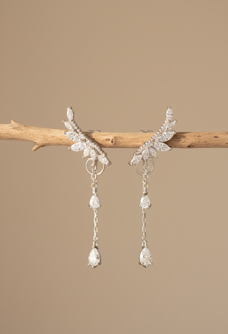 Dione Canada silver leaf drop earrings featuring marquise-cut crystal detailing and elegant dangling stones, displayed on a natural wooden branch in a warm studio setting.