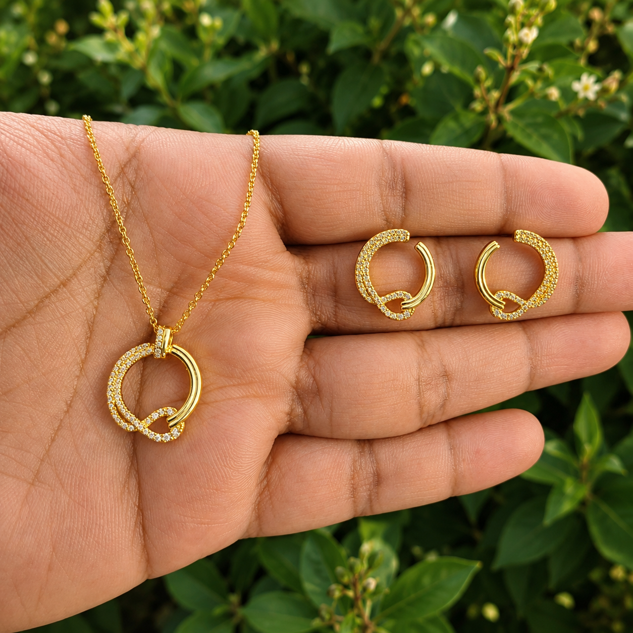 A gold jewelry set consisting of a necklace and matching hoop earrings, displayed on an open hand with a blurred green leafy background.