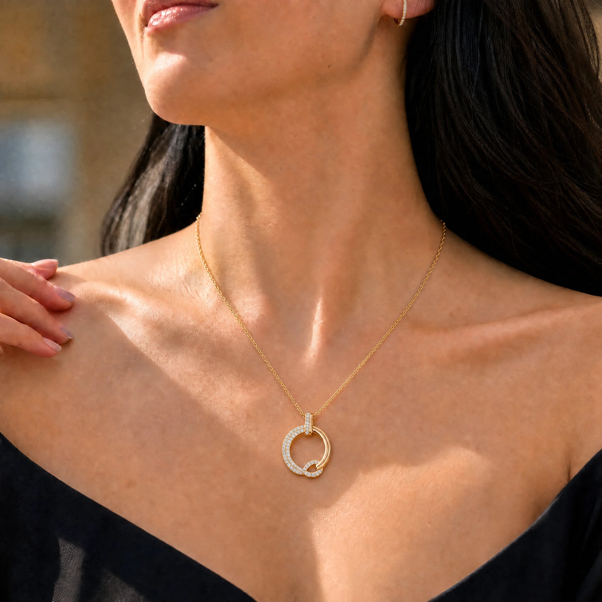 A close-up lifestyle shot of a woman wearing a delicate gold necklace with a circular pendant adorned with small clear stones, set against her bare neck and shoulders.