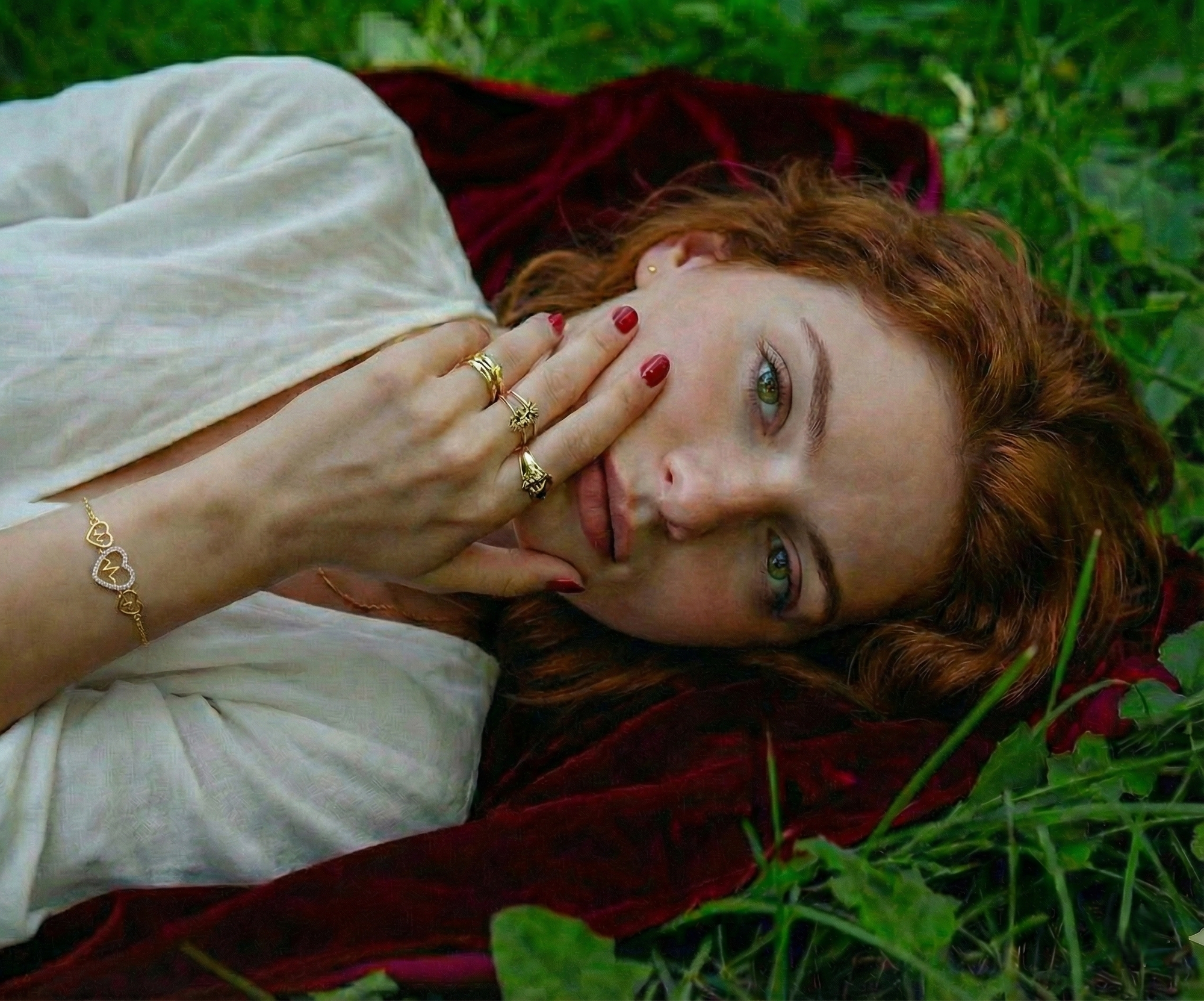 Woman wearing a gold heart bracelet and gold rings during an outdoor photoshoot, lying on grass.