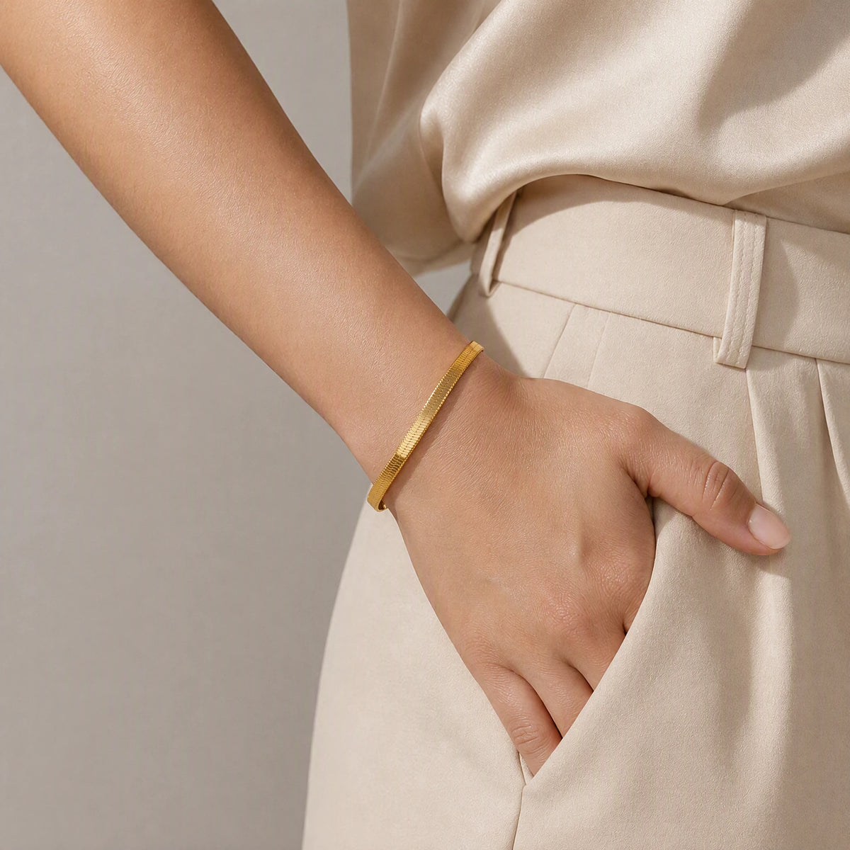 A close-up of a person wearing a slim, polished gold bangle bracelet on their wrist, paired with a beige satin top and matching beige trousers.