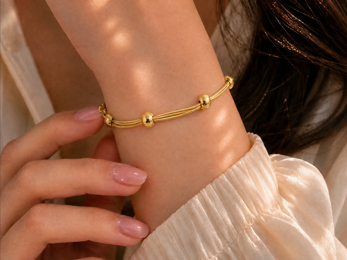 A close-up of a delicate gold bracelet with three small spherical beads worn on a person's wrist, with a hand gently touching the bracelet and a soft cream-colored blouse in the background.