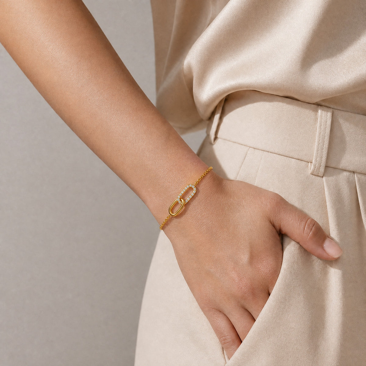 A close-up of a person's hand resting in a pocket, wearing a delicate gold chain bracelet with a double-O ring design set with small clear crystals. The person is wearing a beige satin top and matching trousers against a neutral gray background.