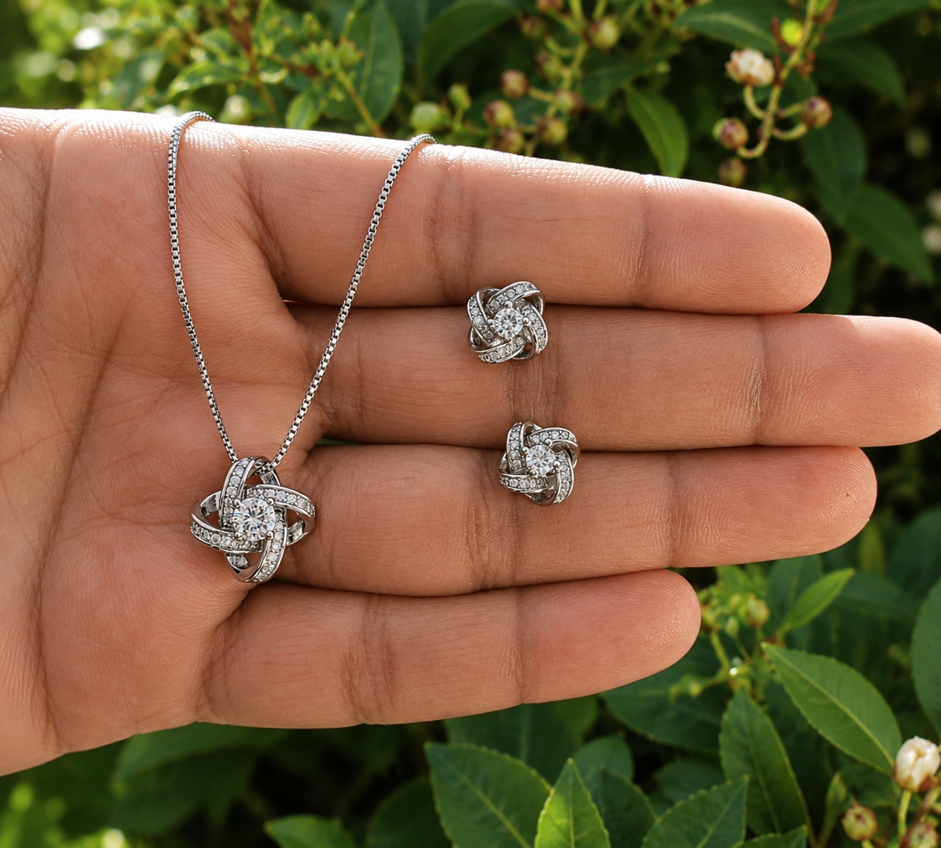 A silver-toned jewelry set featuring a knot pendant necklace and matching stud earrings, displayed on an open hand with a blurred green garden background.