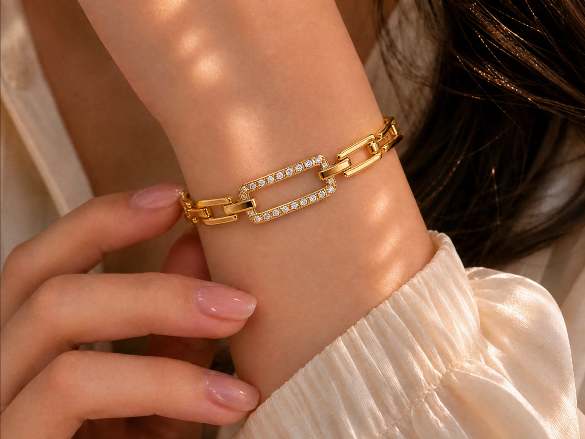 A close-up of a gold chain bracelet with rectangular links, some set with small clear stones, worn on a person's wrist. The person is wearing a cream-colored top, and their hand with pink manicured nails is resting near the bracelet.
