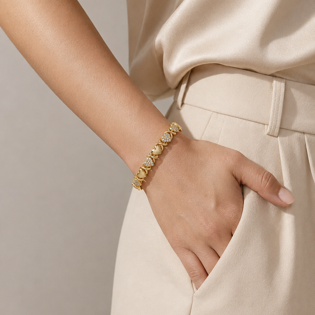 A close-up of a person wearing a gold heart-link bracelet with clear stones, paired with a cream satin top and matching cream trousers.